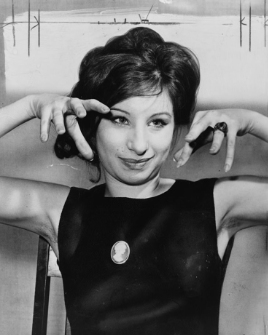 Young woman wearing a dark sleeveless dress with a cameo broach poses with both hands raised in a gesture like animal claws.
