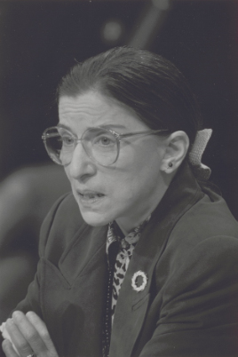 Half-length portrait of a seated woman speaking. She wears a jacket and glasses with her hair pulled back.